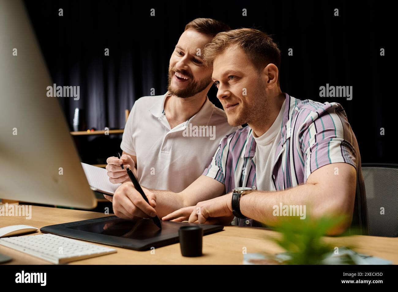 Two men in casual attire, happily collaborating on a project using a laptop at a desk. Stock Photo