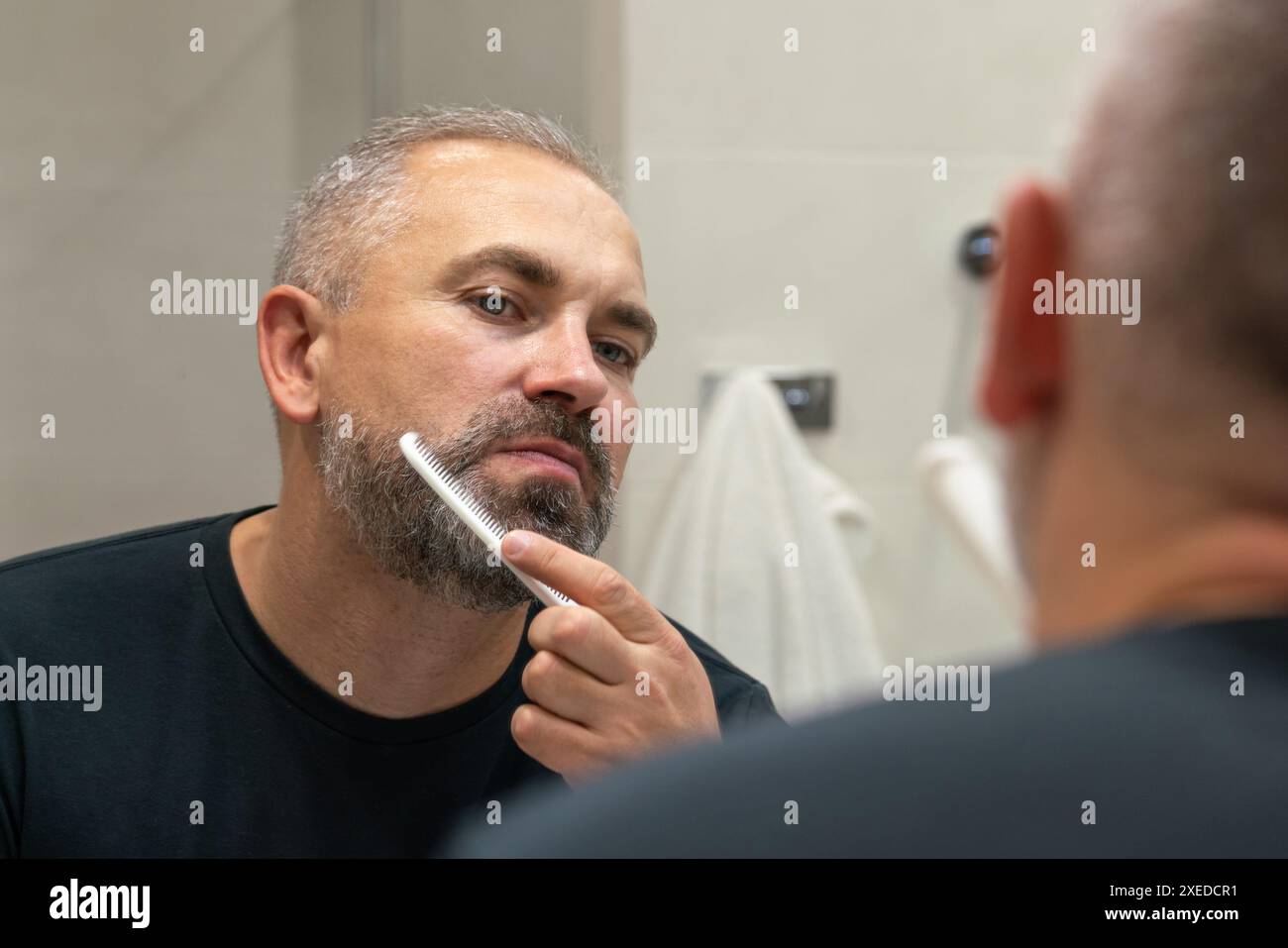 Middle-aged handsome man brushing his beard in the morning in bathroom looking in mirror Stock Photo