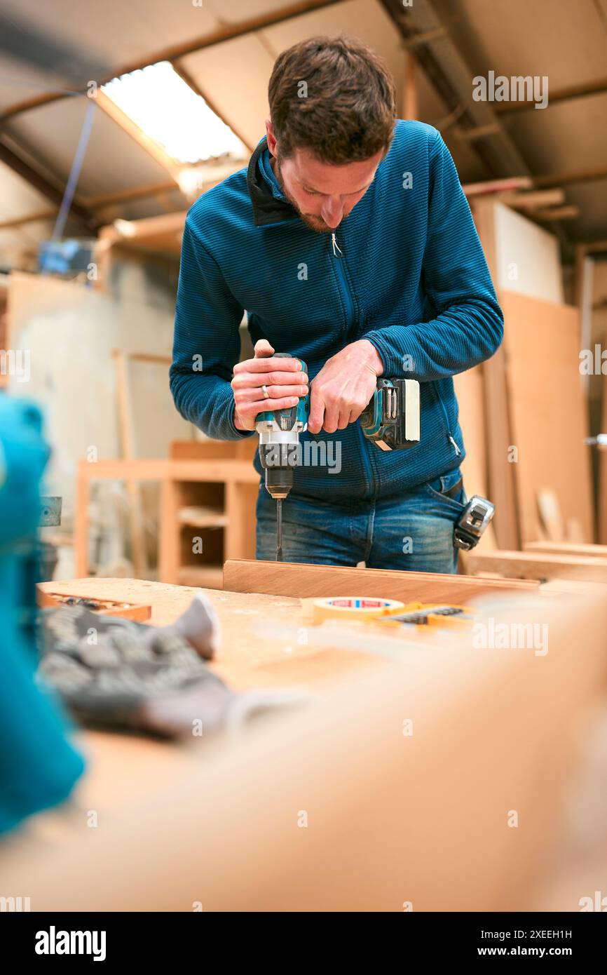Carpenter At Workbench In Workshop Using Cordless Electric Drill Stock Photo