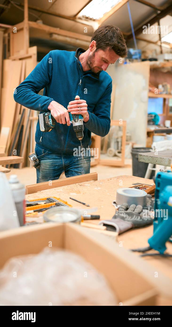 Carpenter At Workbench In Workshop Using Cordless Electric Drill Stock Photo