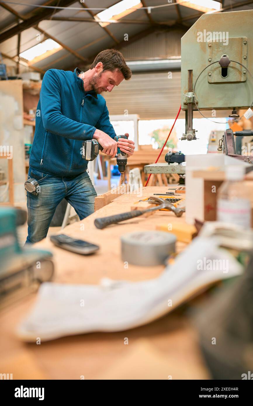 Carpenter At Workbench In Workshop Using Cordless Electric Drill Stock Photo