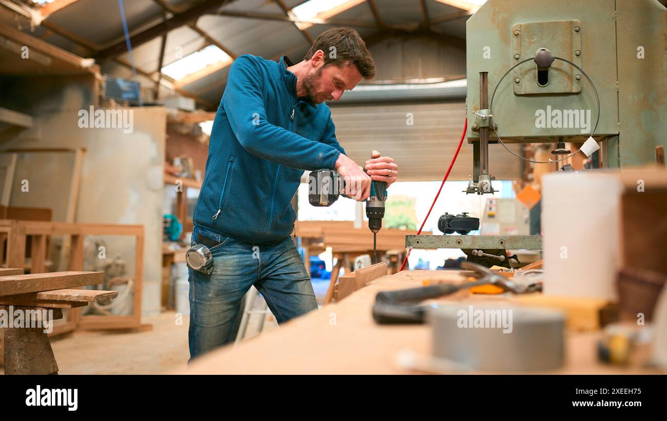 Carpenter At Workbench In Workshop Using Cordless Electric Drill Stock Photo