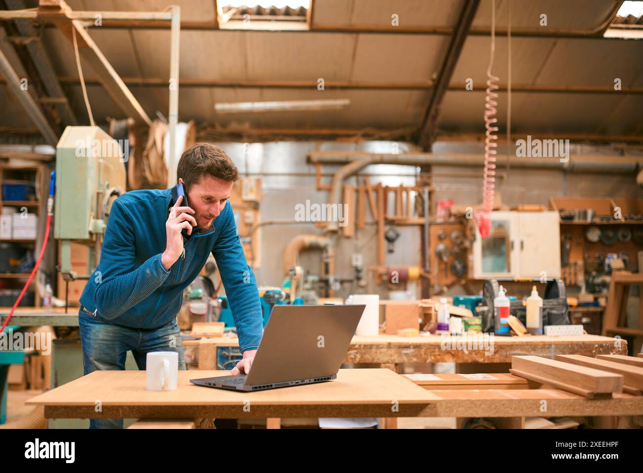Carpenter Working In Woodwork Workshop Talking On Mobile Phone And Using Laptop Stock Photo