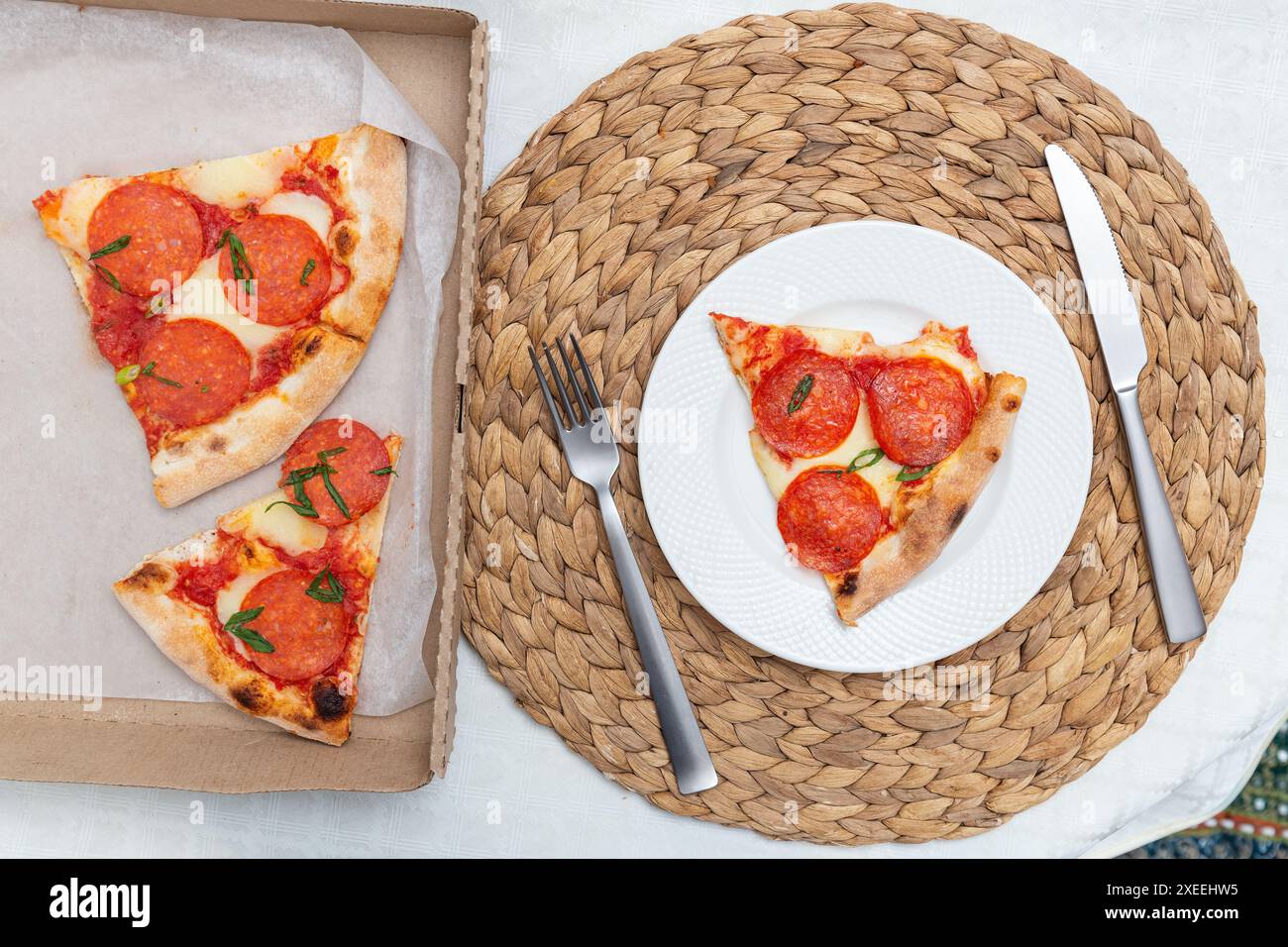 Pepperoni pizza slice on a white plate next to a takeout box with more slices, top view Stock Photo