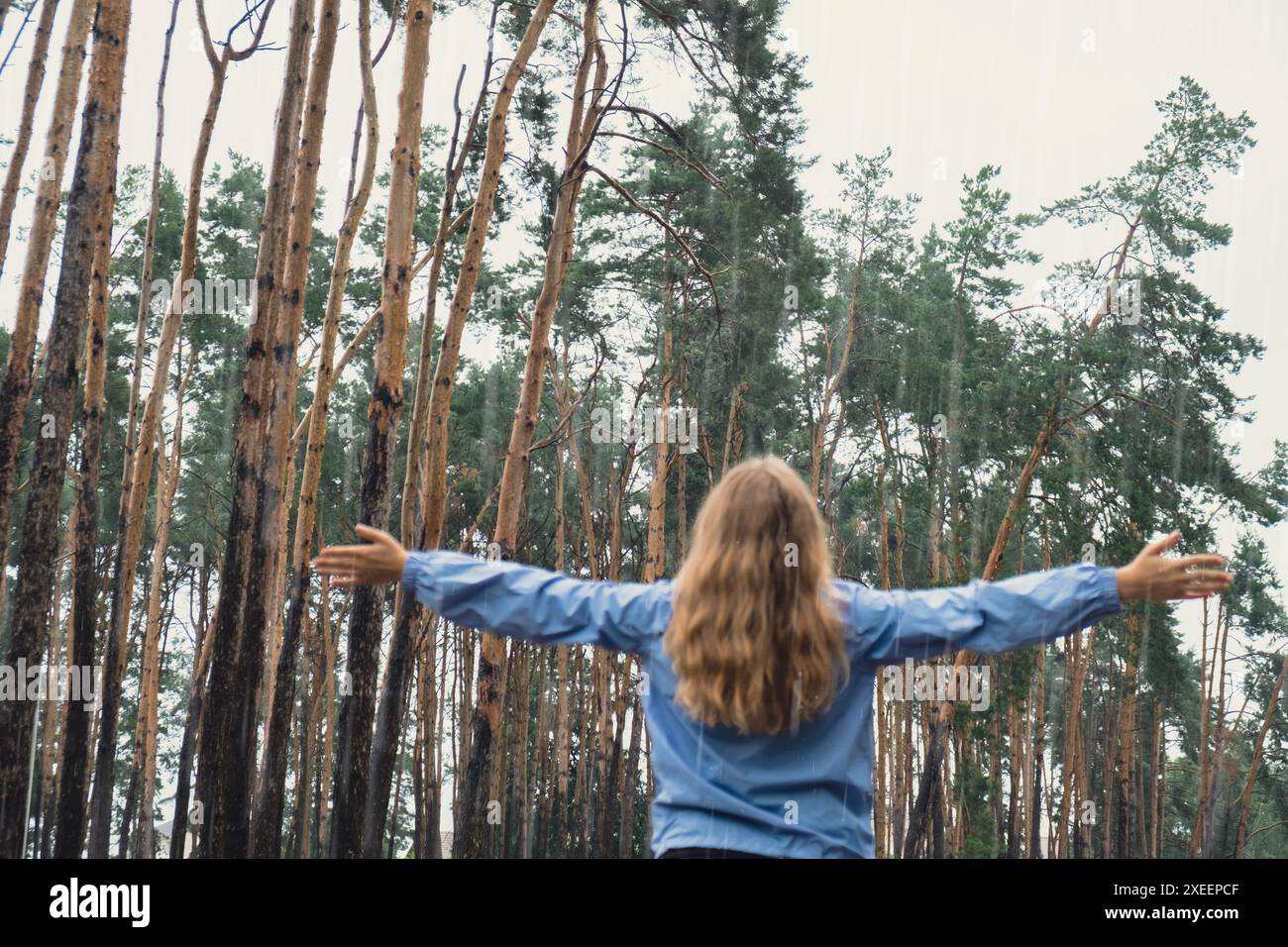 Out of focus Back view young woman walk in blue raincoat enjoying the woods in park. Open arms outdoors in rainy weather forecas Stock Photo