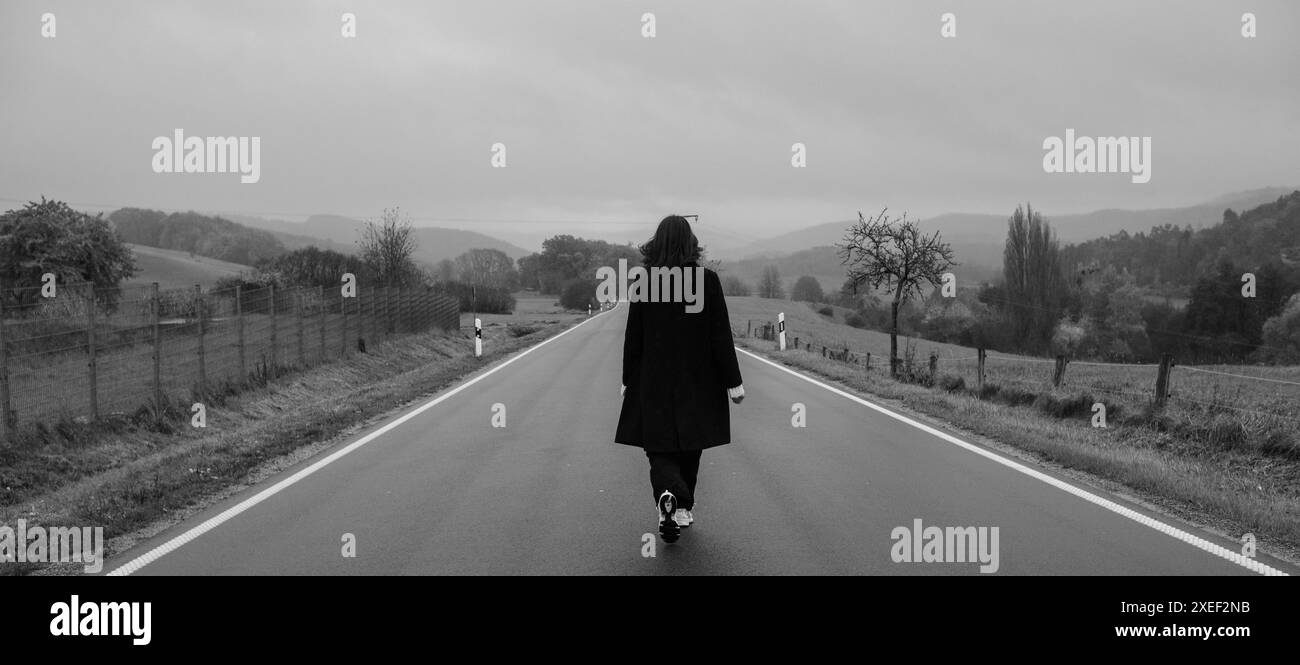 A young woman in a black coat walks alone along a smooth asphalt road, surrounded by yellow trees and grass. Dramatic sky and mountains in the backgro Stock Photo
