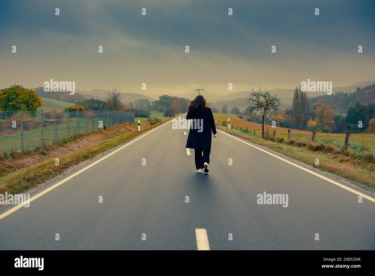 A young woman in a black coat walks alone along a smooth asphalt road, surrounded by yellow trees and grass. Dramatic sky and mountains in the backgro Stock Photo