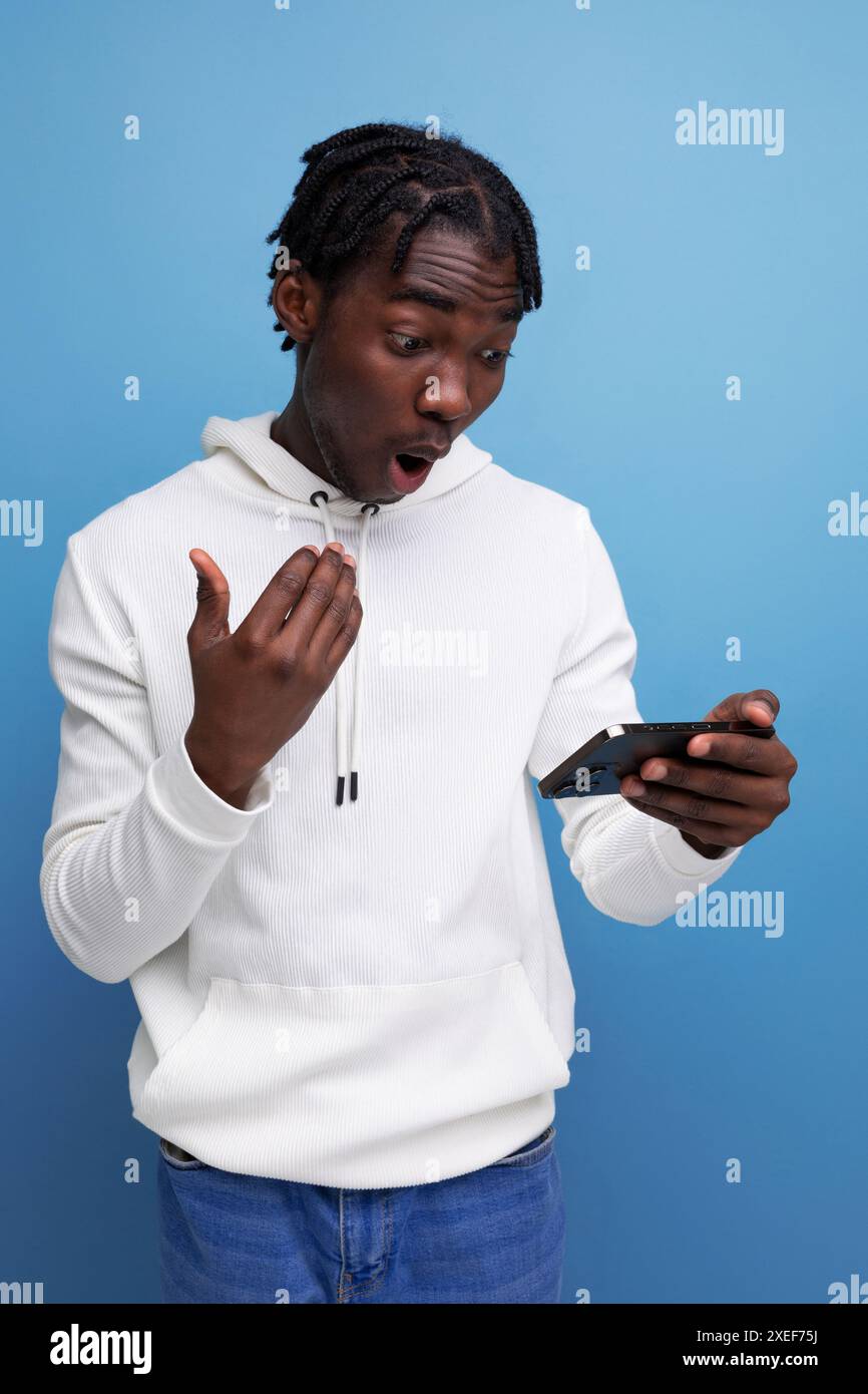 Surprised young african guy in a white sweatshirt understands the smartphone Stock Photo