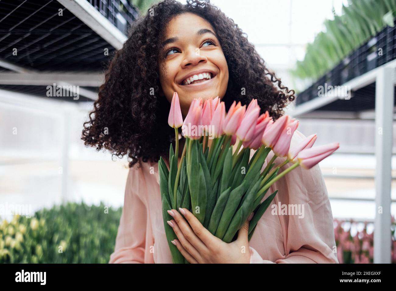 Beautiful romantic smiling african ethnicity girl holding bouquet of pink tulips in greenhouse. Close up of feminine young afro Stock Photo