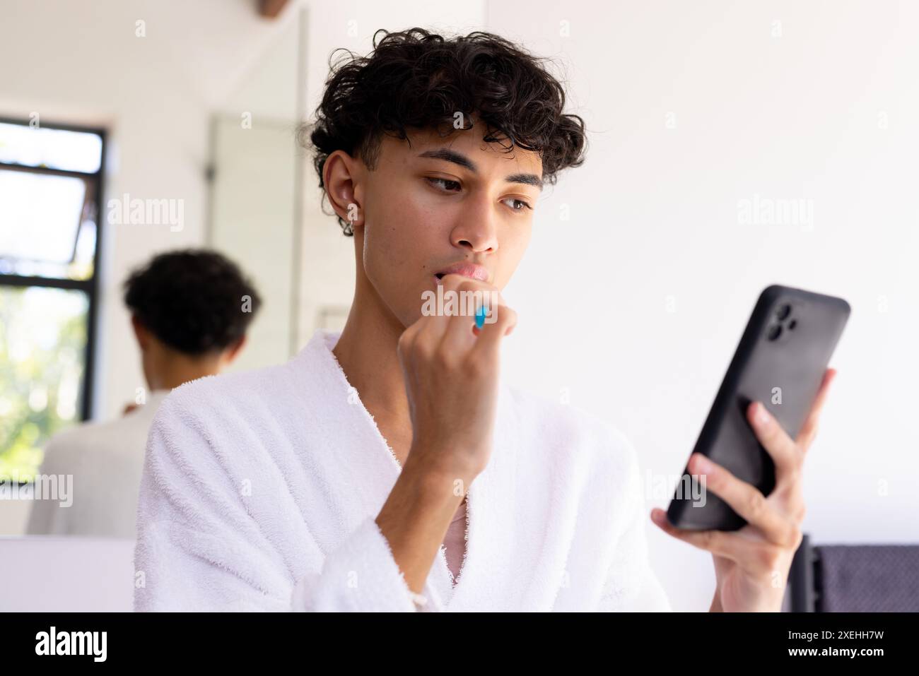 Brushing teeth, young young man in bathrobe using smartphone in bathroom Stock Photo