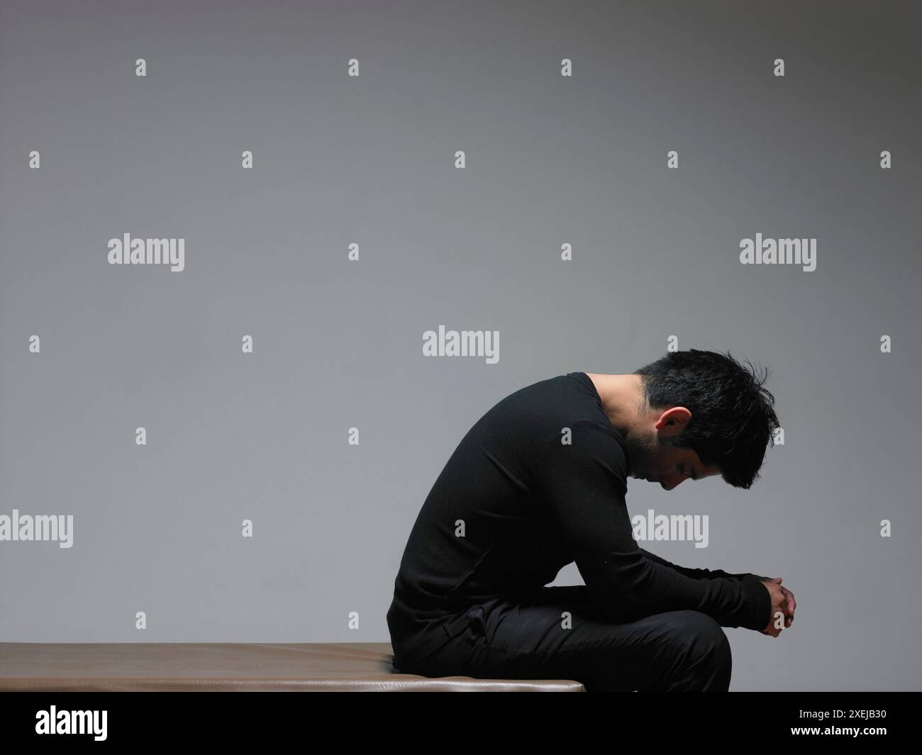 A Hispanic man dressed in black sits with hands clasped and head down. Stock Photo