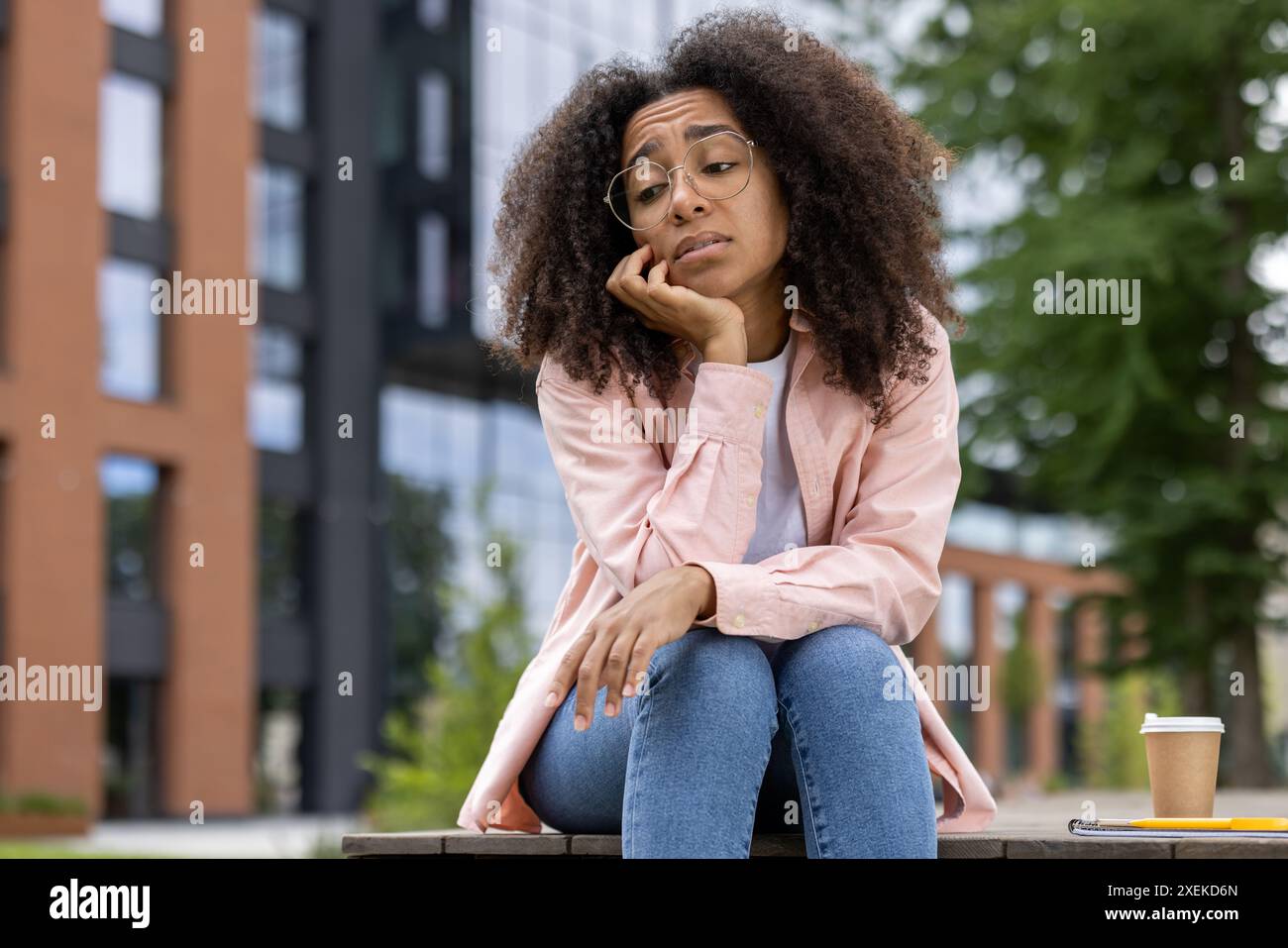 Young woman with curly hair and glasses sitting on bench in urban park, looking bored or sad. Casual attire with a coffee cup and notebook nearby. Stock Photo