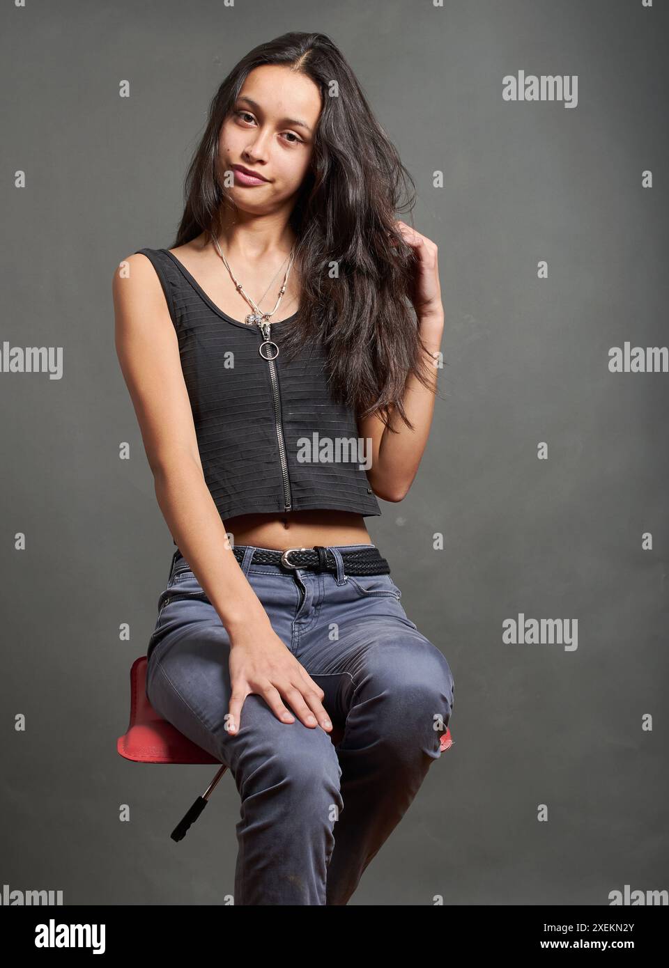 Photograph of a young Hispanic teenage girl sitting posing on a seat looking at the camera. The girl has long black hair and is wearing a casual outfi Stock Photo