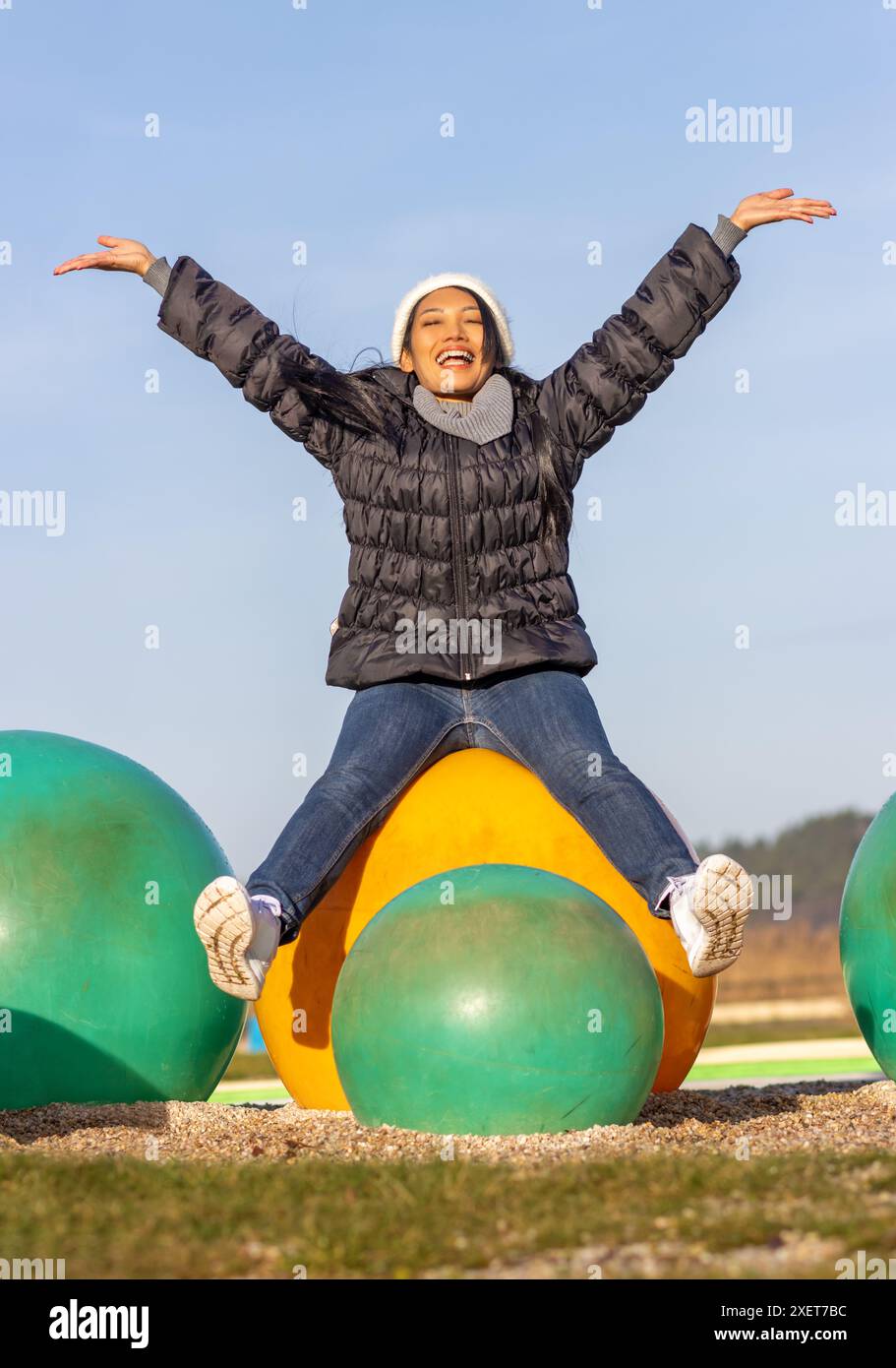 A cheerful woman is sitting on a large colorful ball on the playground Stock Photo