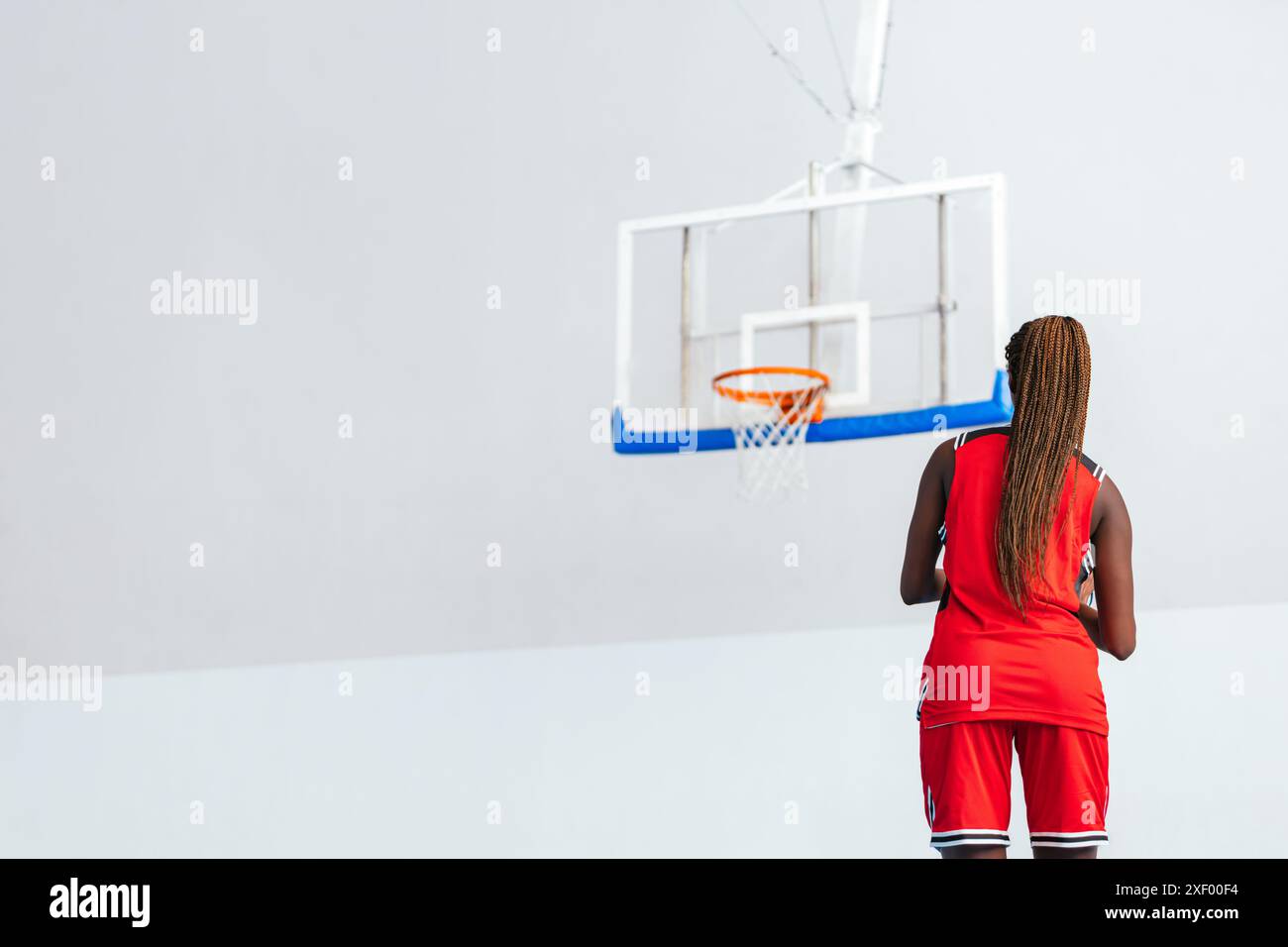 Horizontal photo a female basketball player in a red uniform stands on an indoor court, holding a basketball and focusing on the hoop, preparing for h Stock Photo