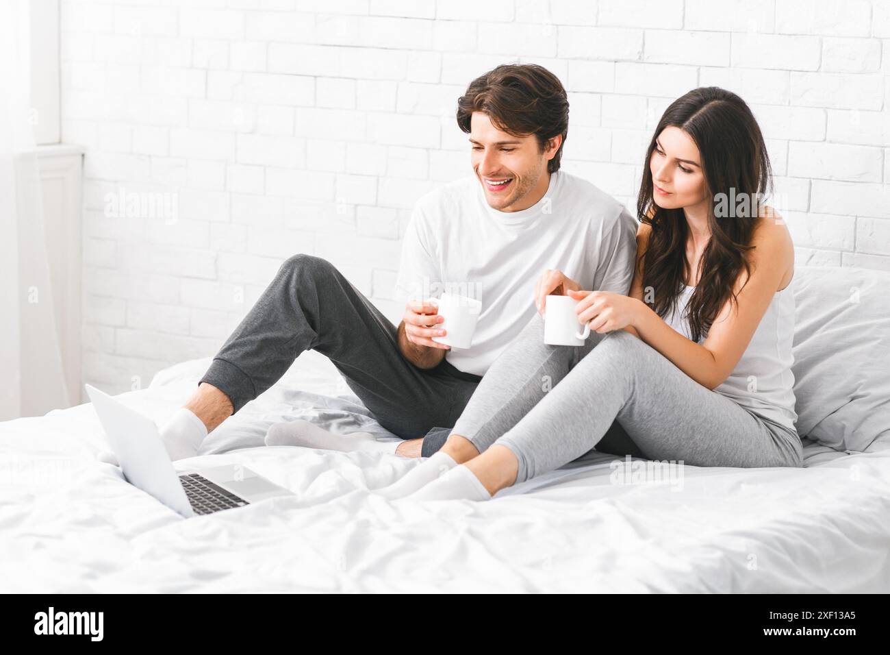 Couple Sitting On Bed Drinking Coffee And Using Laptop Stock Photo