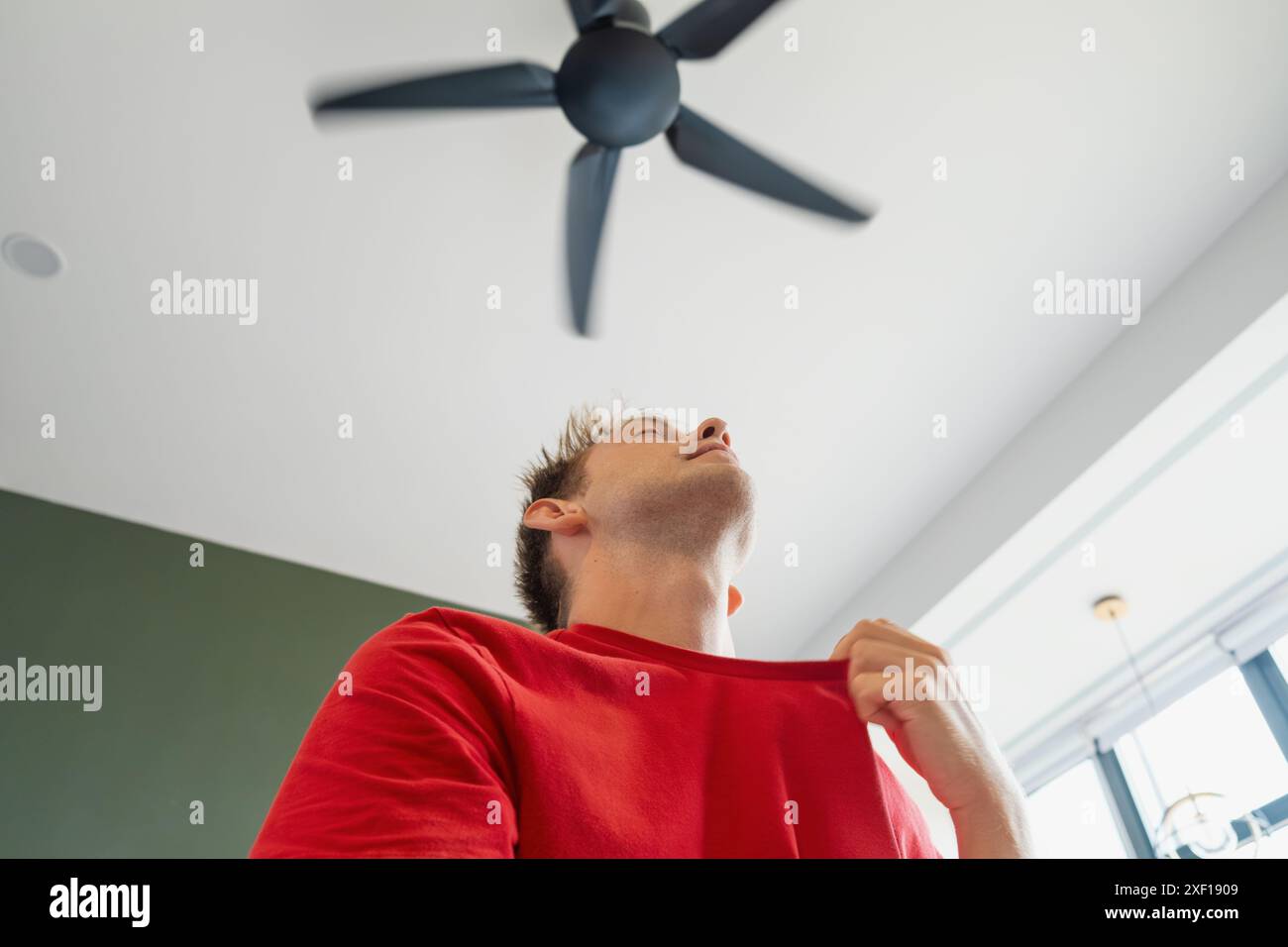 Man sitting under ceiling fan at home in living room, cooling off during hot summer weather, suffering from heat, high temperature. Sweaty exhausted Stock Photo