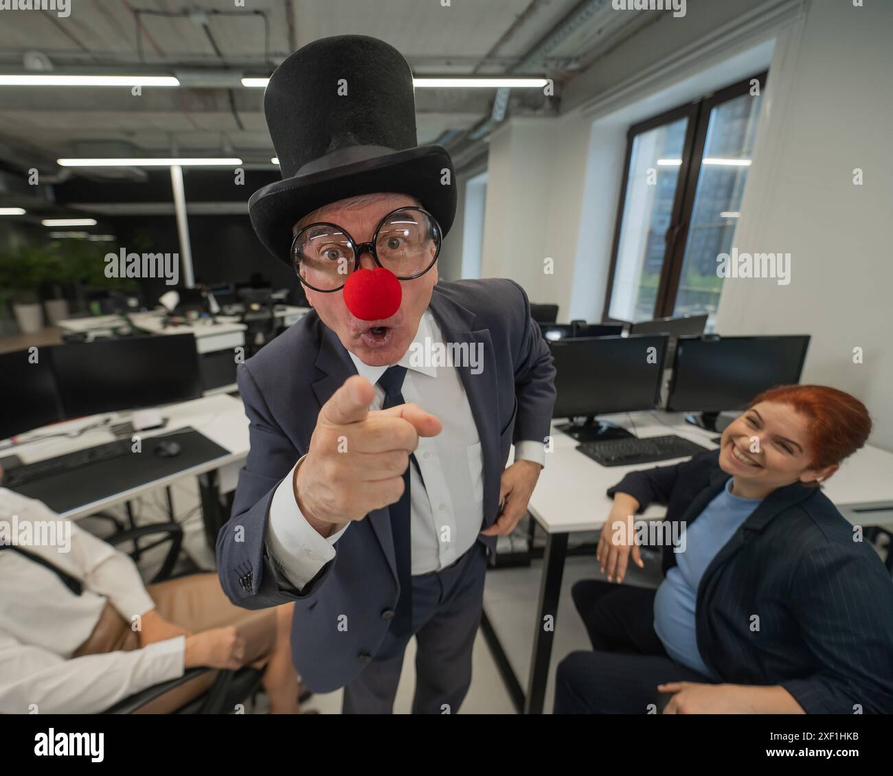 An elderly Caucasian man in a clown costume amuses two Caucasian women in the office.  Stock Photo