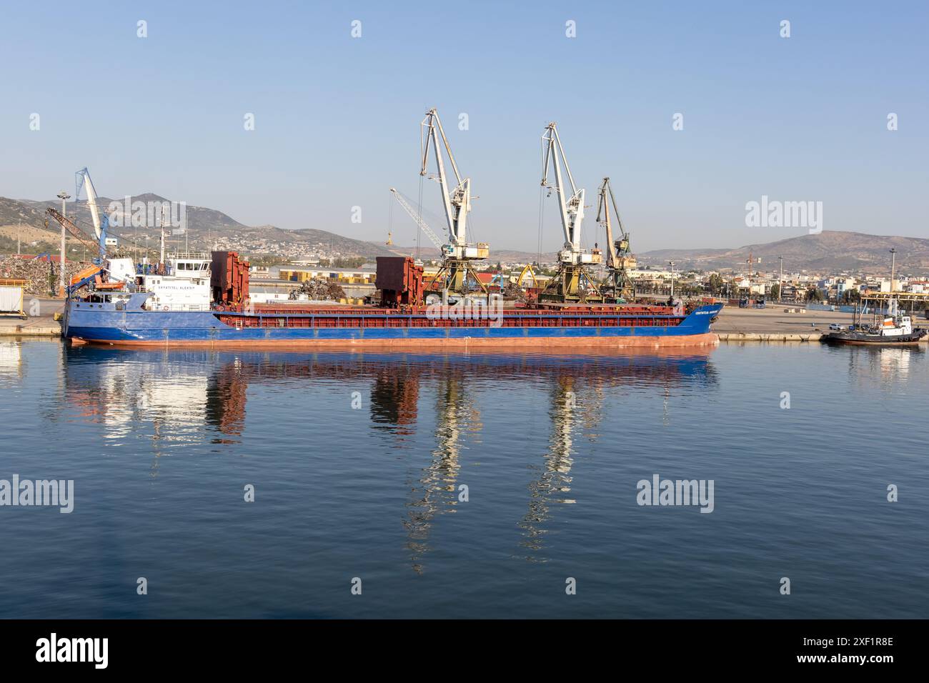 A large freight ship docked at a busy industrial port with cranes for cargo handling, and a cityscape in the background. Stock Photo
