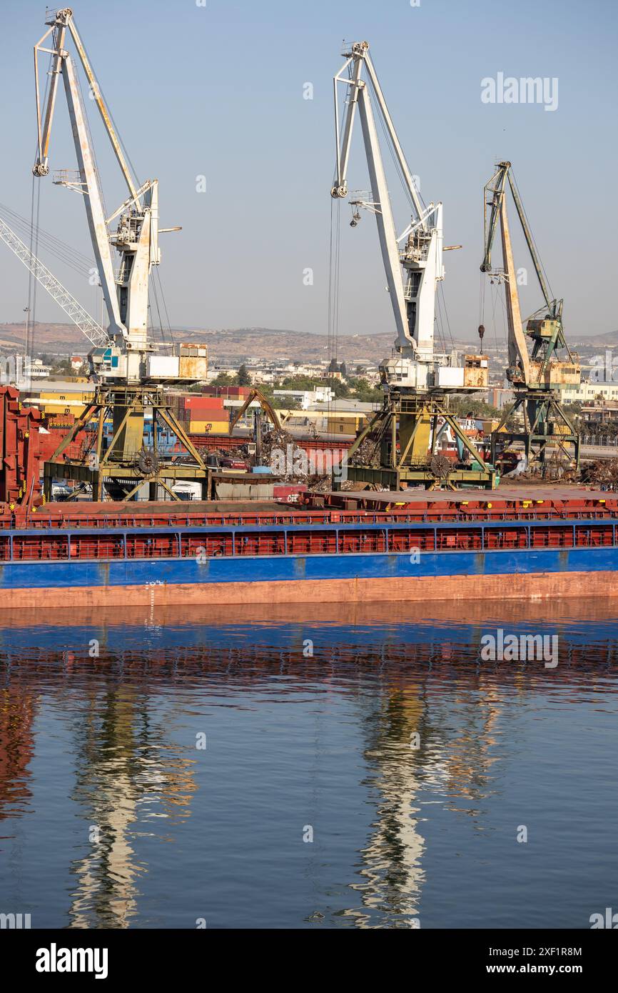 A large freight ship docked at a busy industrial port with cranes for cargo handling, and a cityscape in the background. Stock Photo