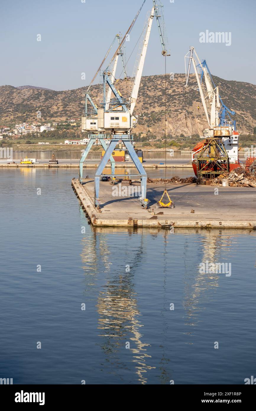 A large freight ship docked at a busy industrial port with cranes for cargo handling, and a cityscape in the background. Stock Photo