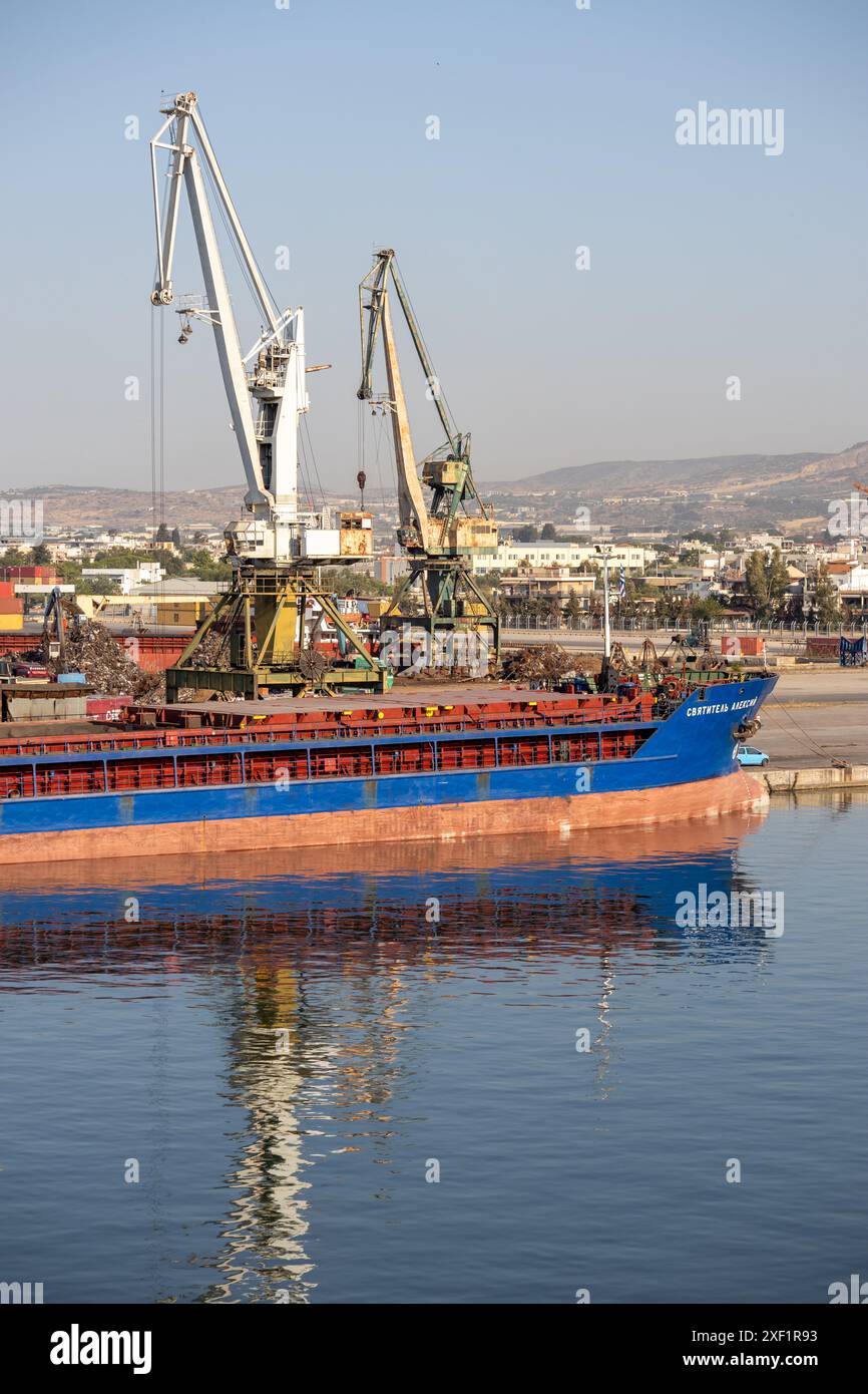 A large freight ship docked at a busy industrial port with cranes for cargo handling, and a cityscape in the background. Stock Photo