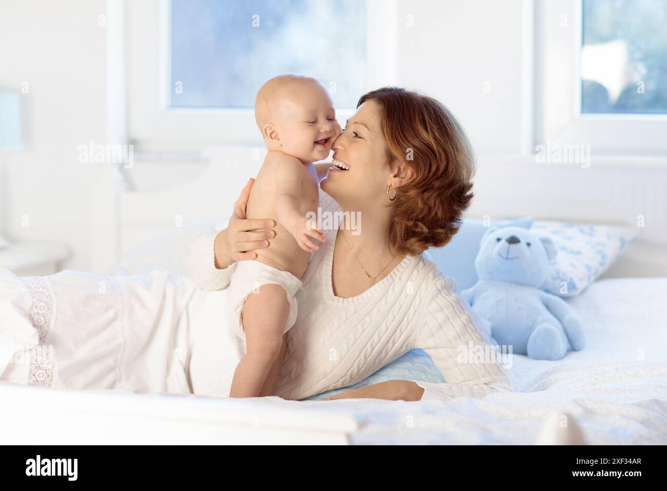 Mother and child on a white bed. Mom and baby boy in diaper playing in sunny bedroom. Parent and little kid relaxing at home. Stock Photo