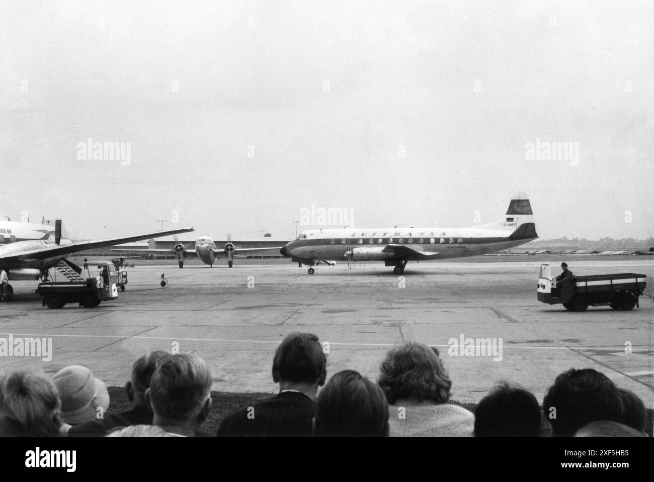 Germany Circa. 1960 – A group of spectators watching aircraft at an airport. In the foreground is the Vickers Viscount 814 turboprop airliner of the German airline, Deutsche Lufthansa; registration, D-ANEF. The aircraft is decorated in its original livery. This aircraft was in service with Lufthansa from March 1959 until 20 January 1972, when it was purchased by Airwork Services Ltd. Eight days later, the aircraft was damaged beyond repair during the delivery flight, after it made a heavy landing at Hurn Airport, Bournemouth, England. An airport worker is driving a MIAG Plattenwagen vehicle. Stock Photo