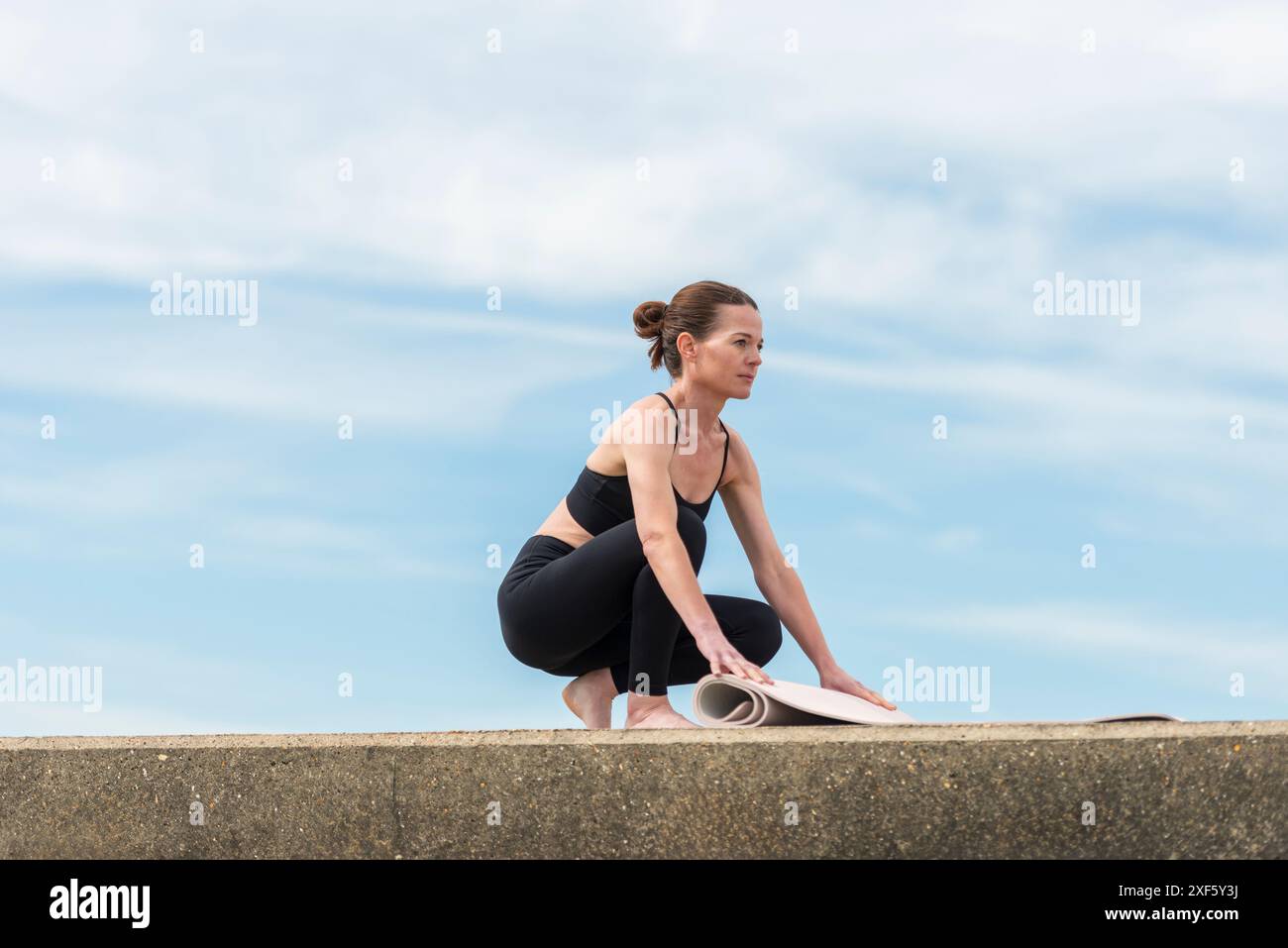 Woman rolling out her yoga mat, outdoors with blue sky background Stock Photo