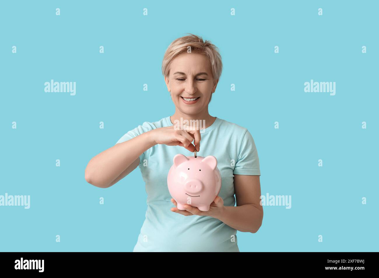 Beautiful mature woman putting coins into pink piggy bank on blue ...