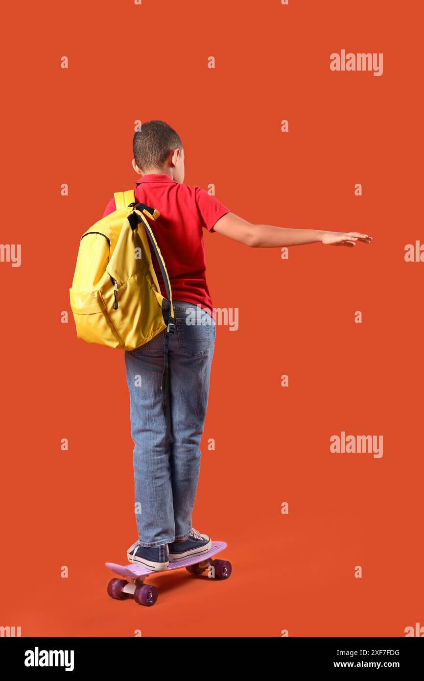 Little schoolboy with backpack skating on orange background, back view ...