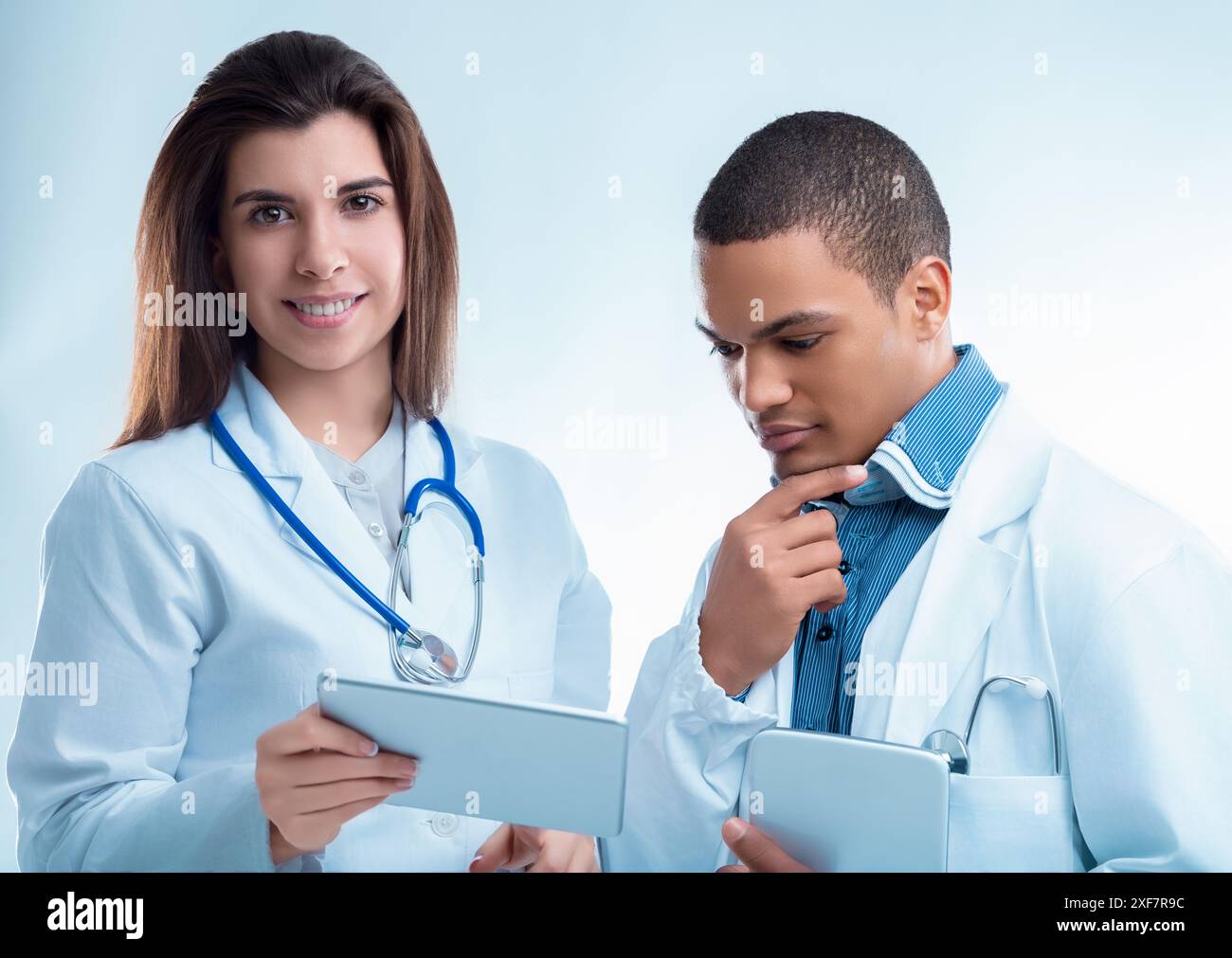 Two young doctors are using digital tablets while wearing white coats and stethoscopes. They are concentrating on the information on the screen Stock Photo