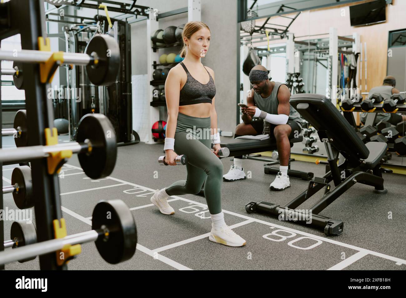 Newbie doing barbell lunge while her African American male coach sitting on bench and writing her training routine in phone Stock Photo