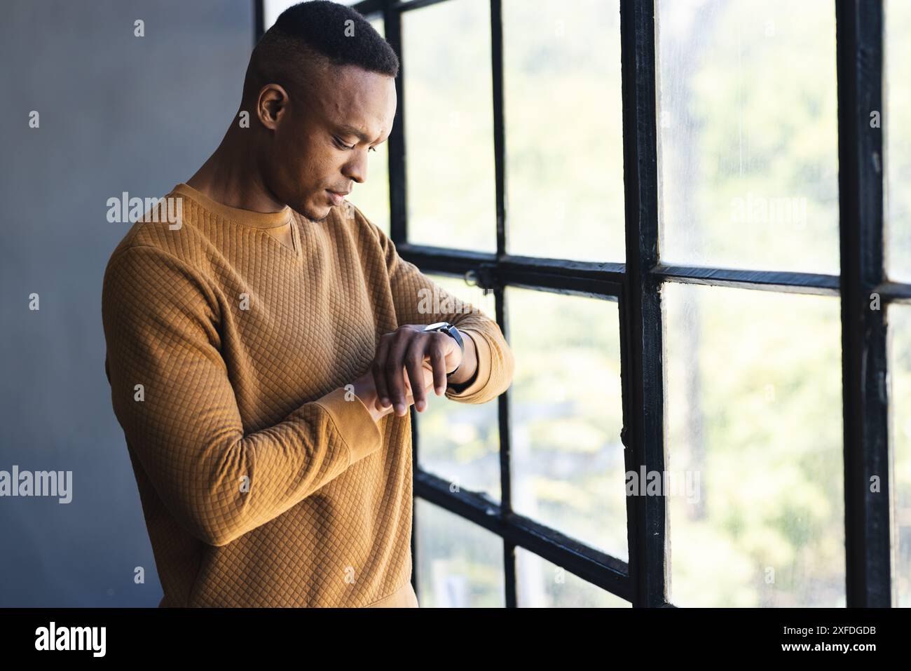 Checking smartwatch, man standing by window in casual sweater, focusing on time Stock Photo