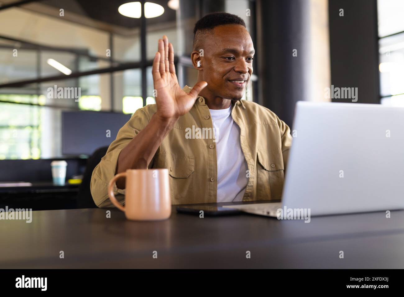 Waving hand, man using laptop and wearing earbuds in modern office ...