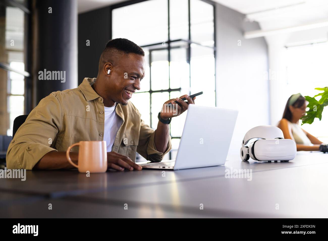 Smiling man using smartphone and laptop in modern office with coffee mug Stock Photo