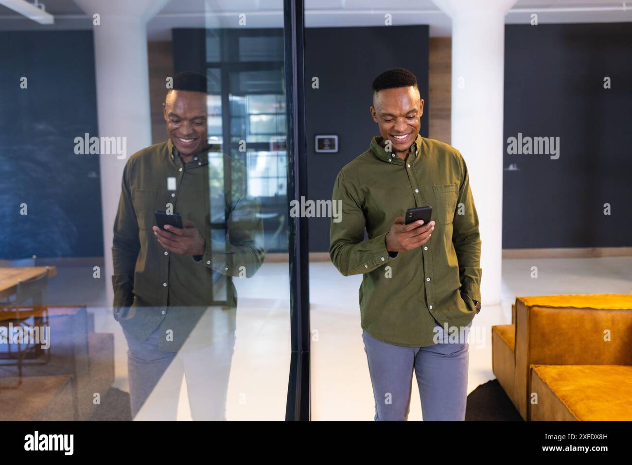 Smiling businessman using smartphone while standing by glass wall in modern office, copy space Stock Photo
