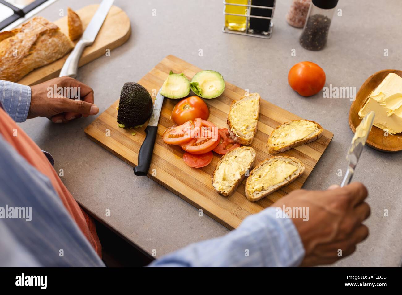 Preparing breakfast, man spreading butter on bread with avocado and tomato slices Stock Photo