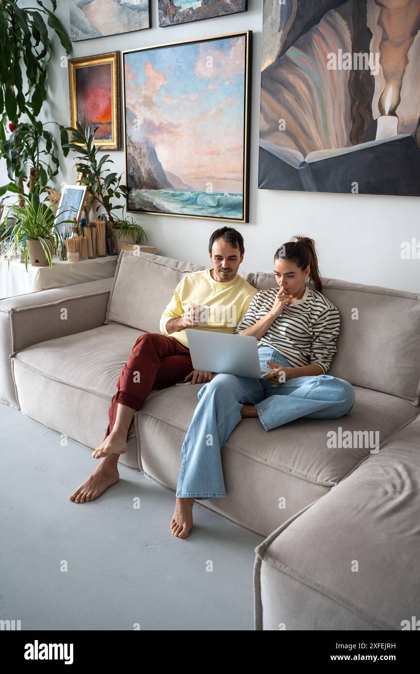Pleased married couple sitting on sofa with laptop at art home studio, spending weekend together. Stock Photo
