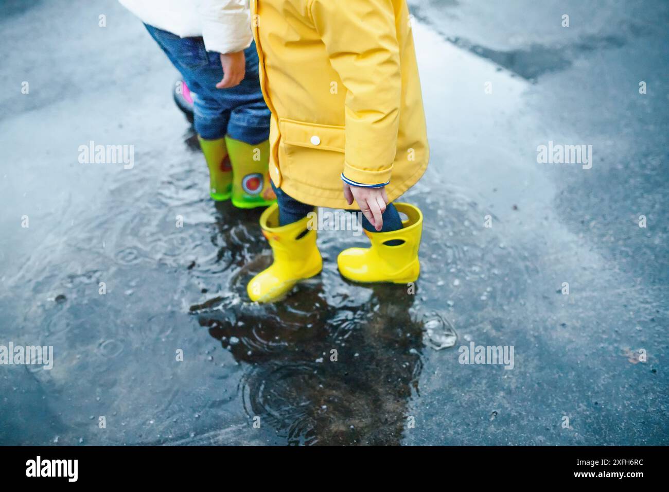 Two children wearing yellow rain boots splash in a puddle after a rain ...