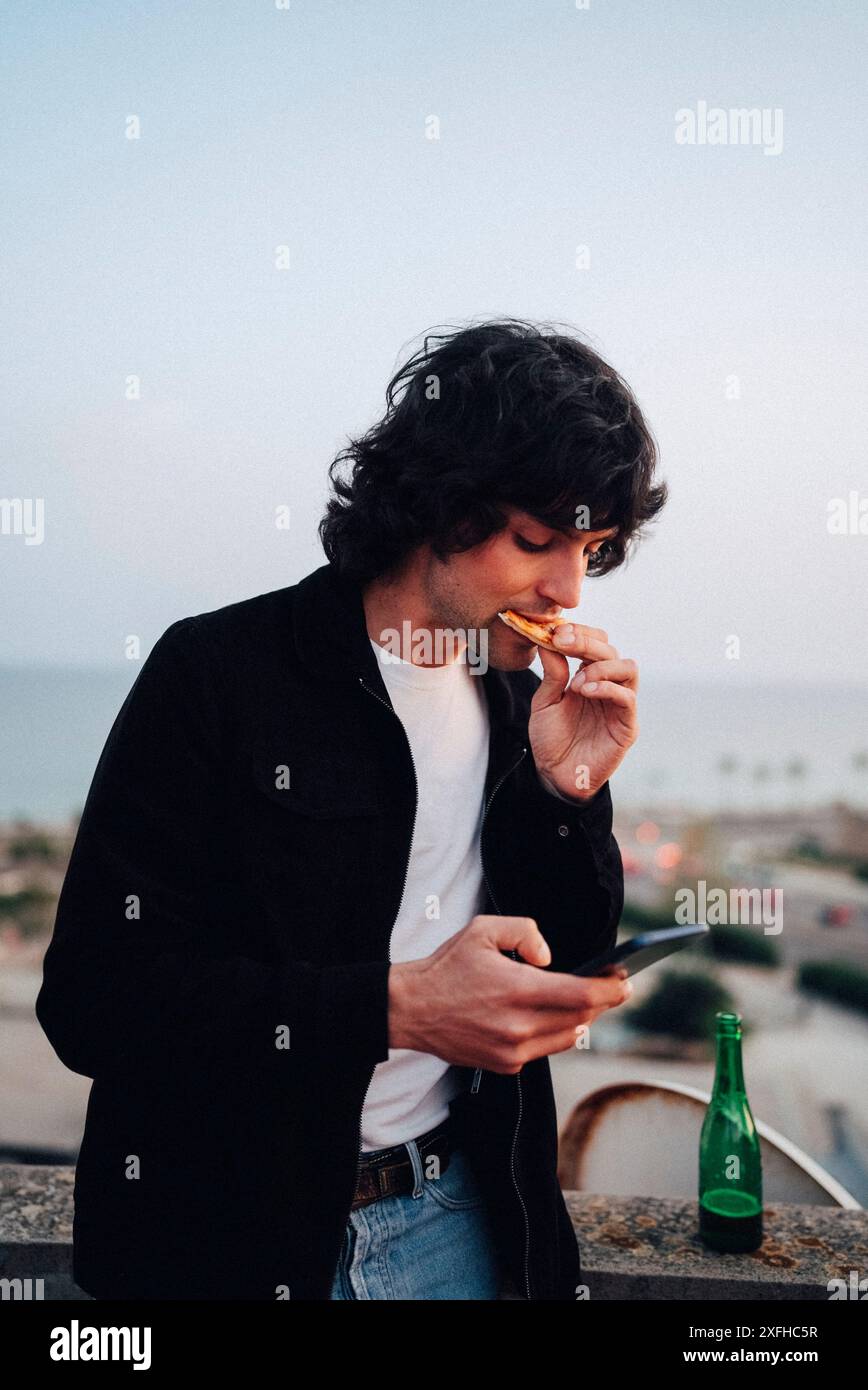 Young man eating pizza and using smart phone standing at rooftop Stock Photo