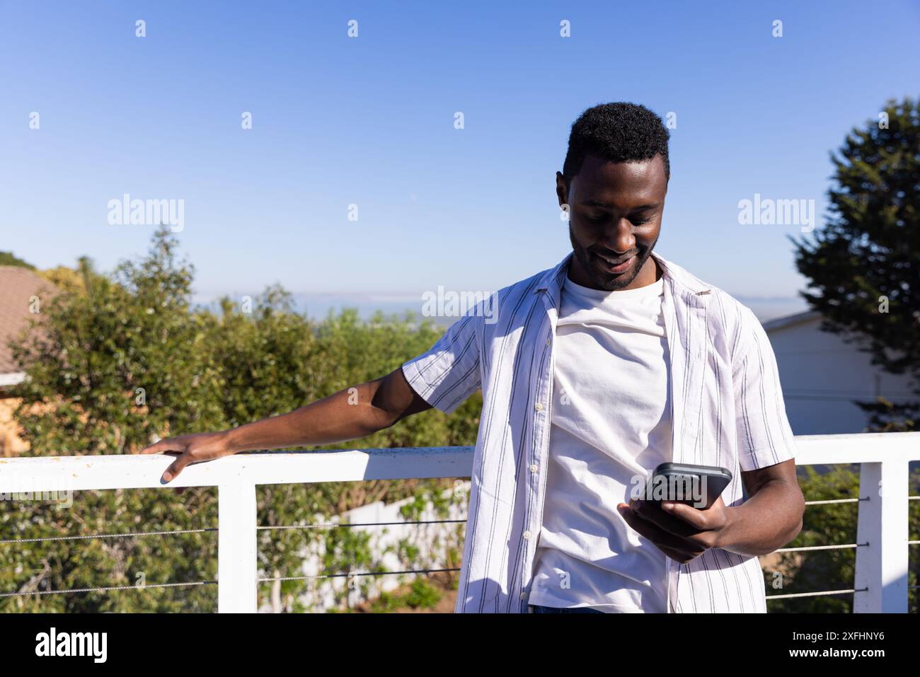 Smiling man using smartphone while standing on balcony, enjoying outdoor view Stock Photo
