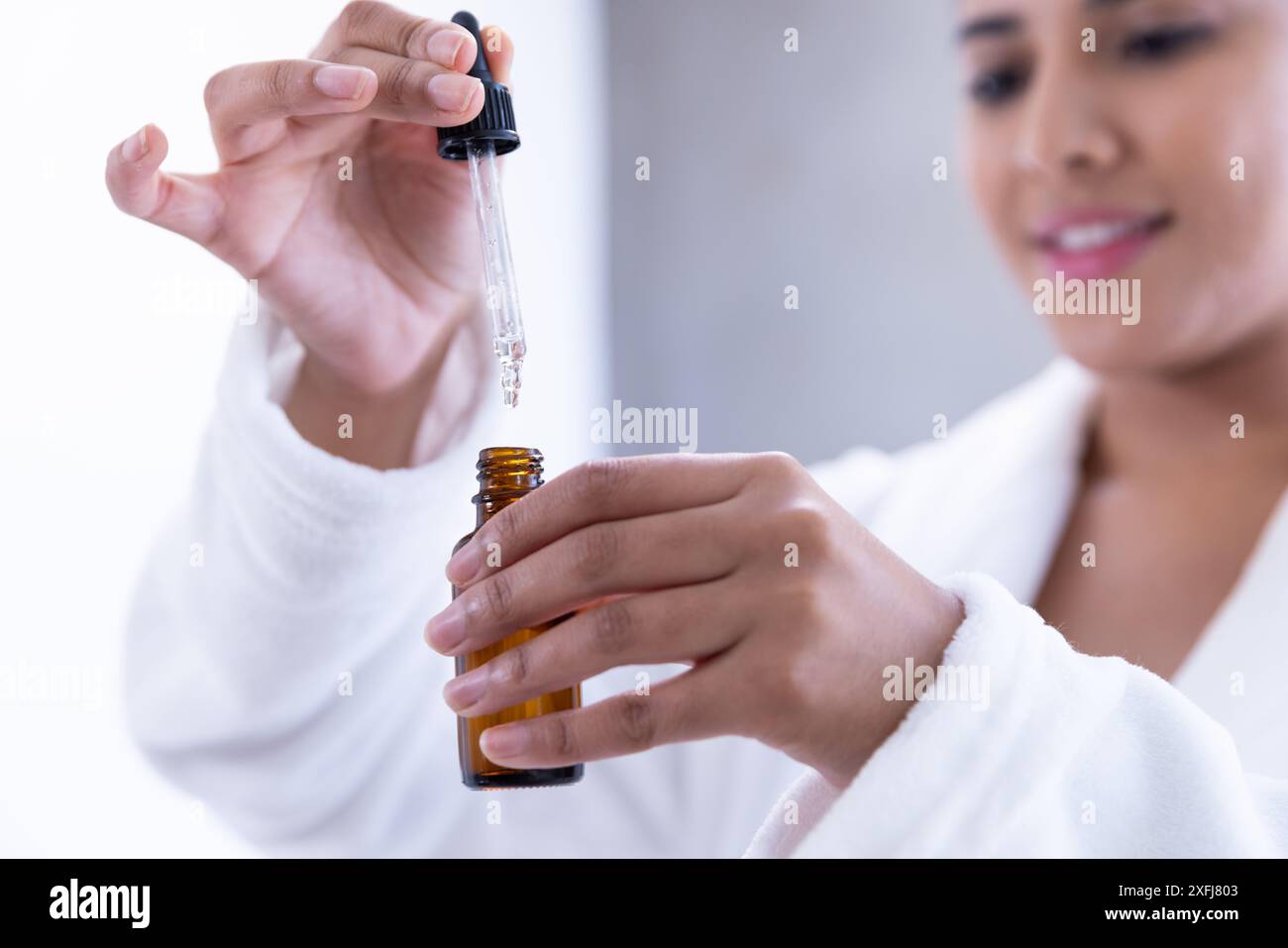 Using dropper bottle, woman in bathrobe applying skincare serum in spa ...