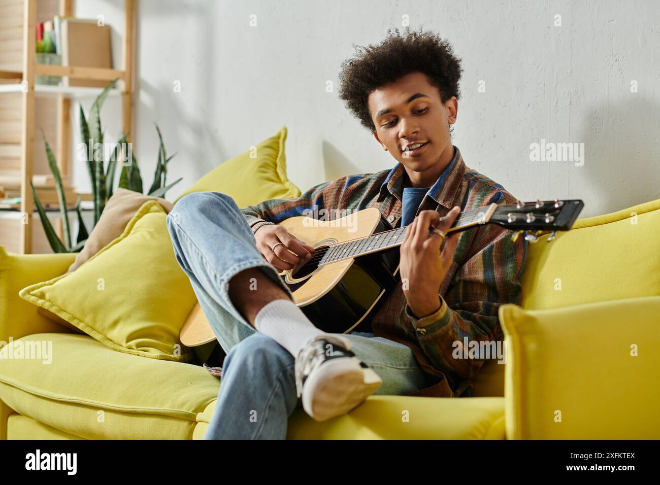 A young African American man playing an acoustic guitar on a yellow couch while talking on his phone. Stock Photo