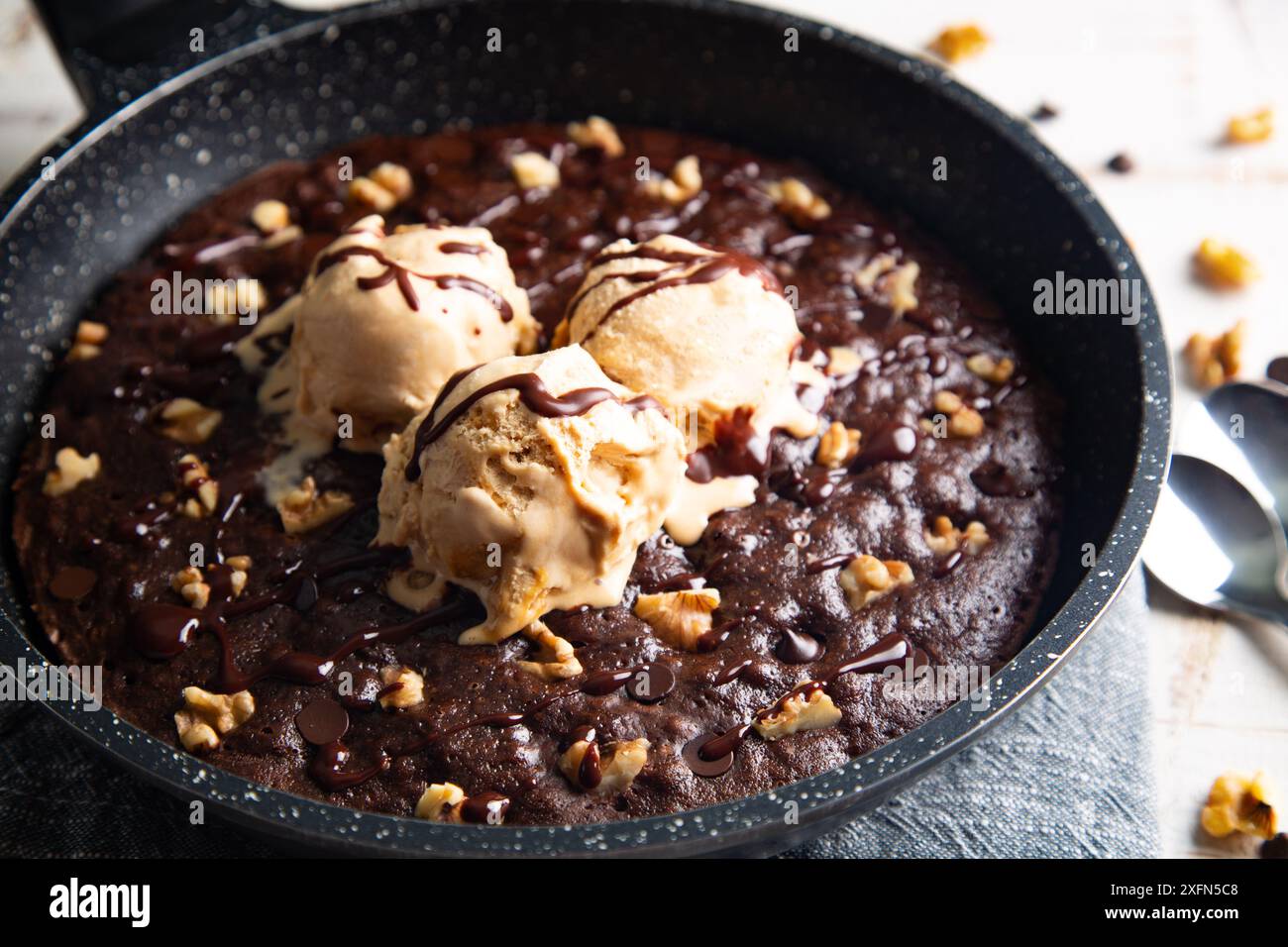 Homemade brownie cooked in a skillet, not baked, with melting vanilla ice cream scoops, nuts, and chocolate chips on a white table Stock Photo