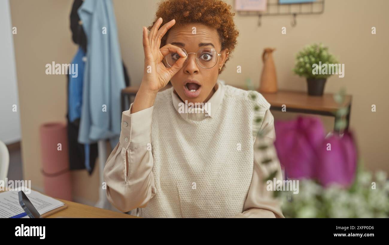 Surprised black woman making an ok gesture over her eye indoors at home ...