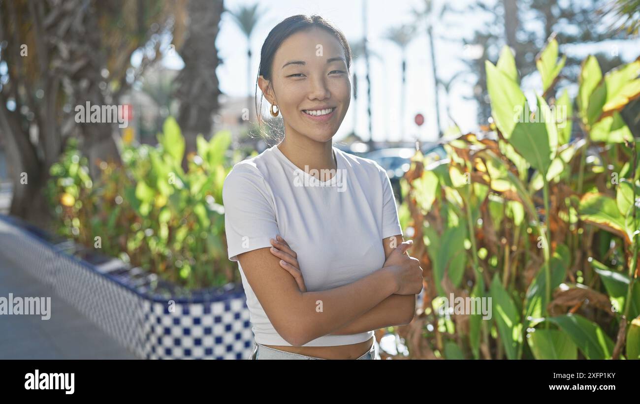 A young asian woman stands arms-crossed, smiling in a sunny, green ...