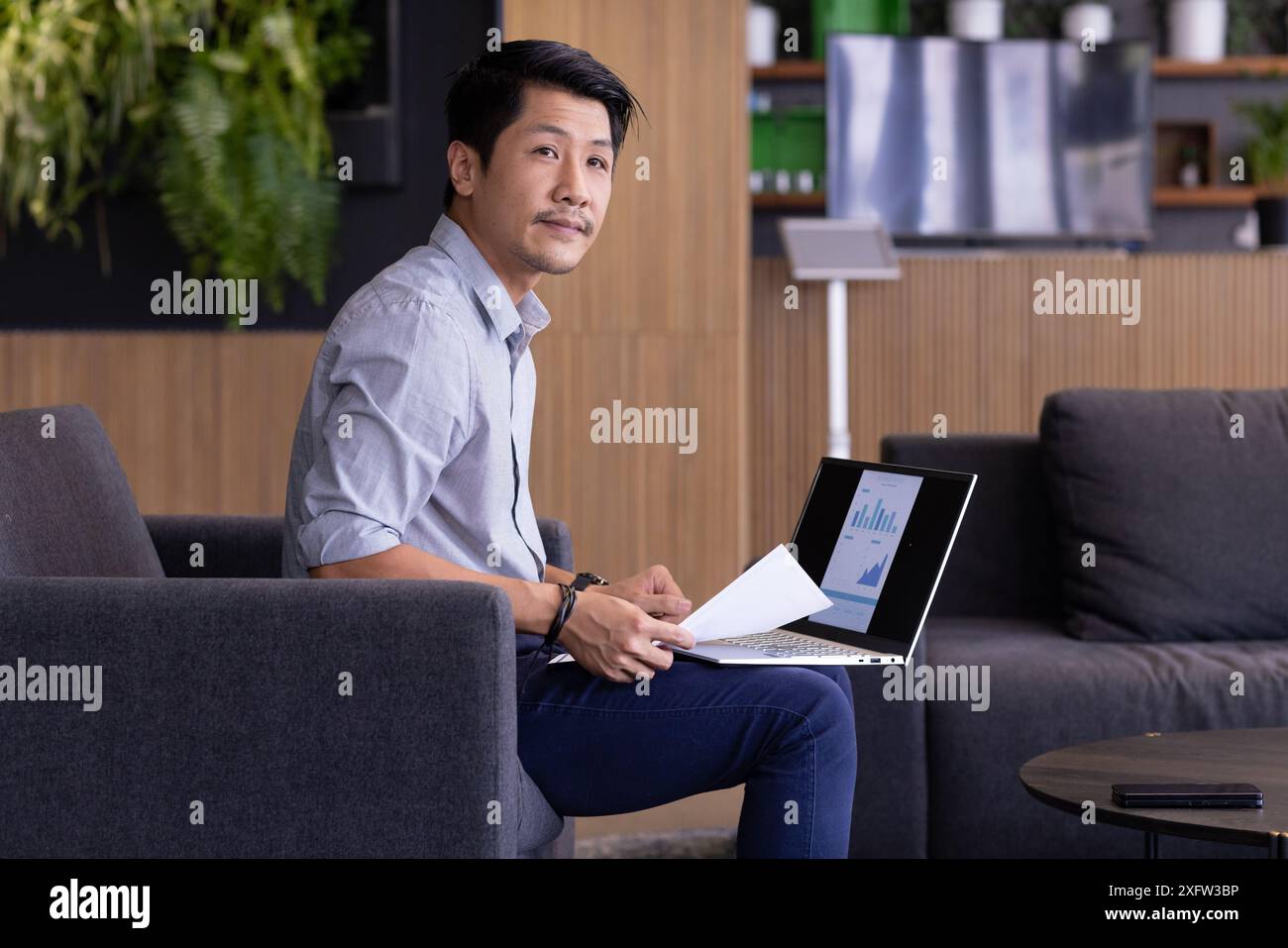 Working on laptop, man analyzing financial charts and holding documents in office Stock Photo
