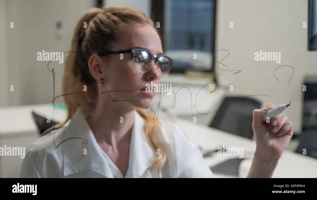 A Caucasian woman in a medical gown thinks and finalizes formulas on a transparent wall.  Stock Photo