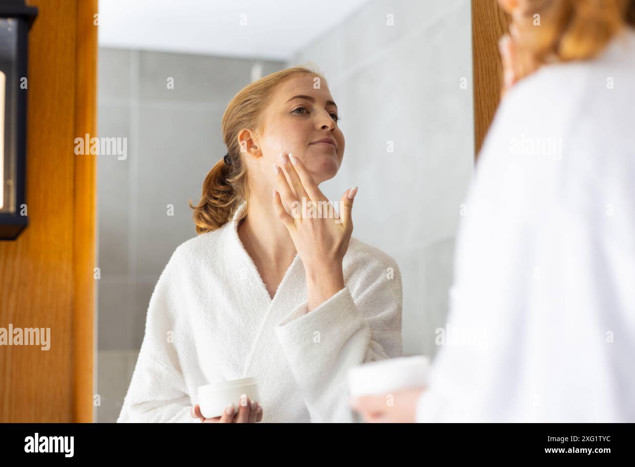 Bathrobe Routine Applying face cream, woman in bathrobe looking in bathroom mirror ...